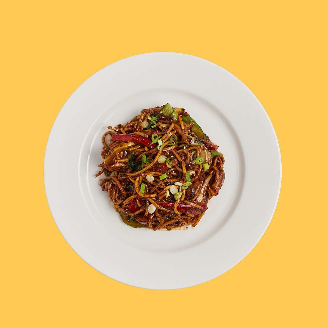 Stir-fried noodles rest piled on a white plate, glistening in dark sauce with peppers and scallions; centered presentation against a solid yellow background.