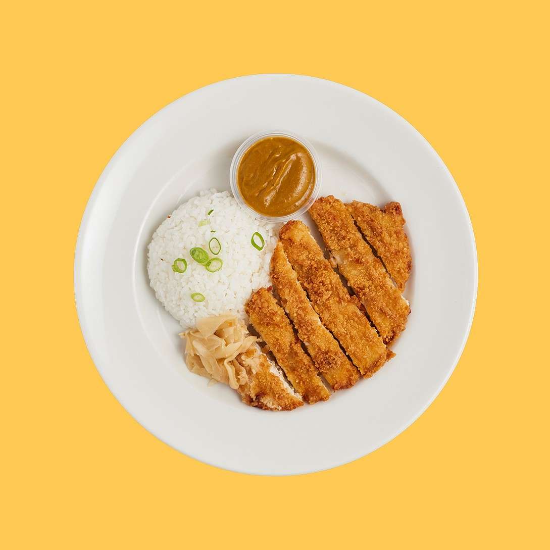 Crispy breaded cutlet slices beside white rice topped with scallions, a cup of brown curry sauce, and pickled ginger, arranged on a white plate against a solid yellow background.