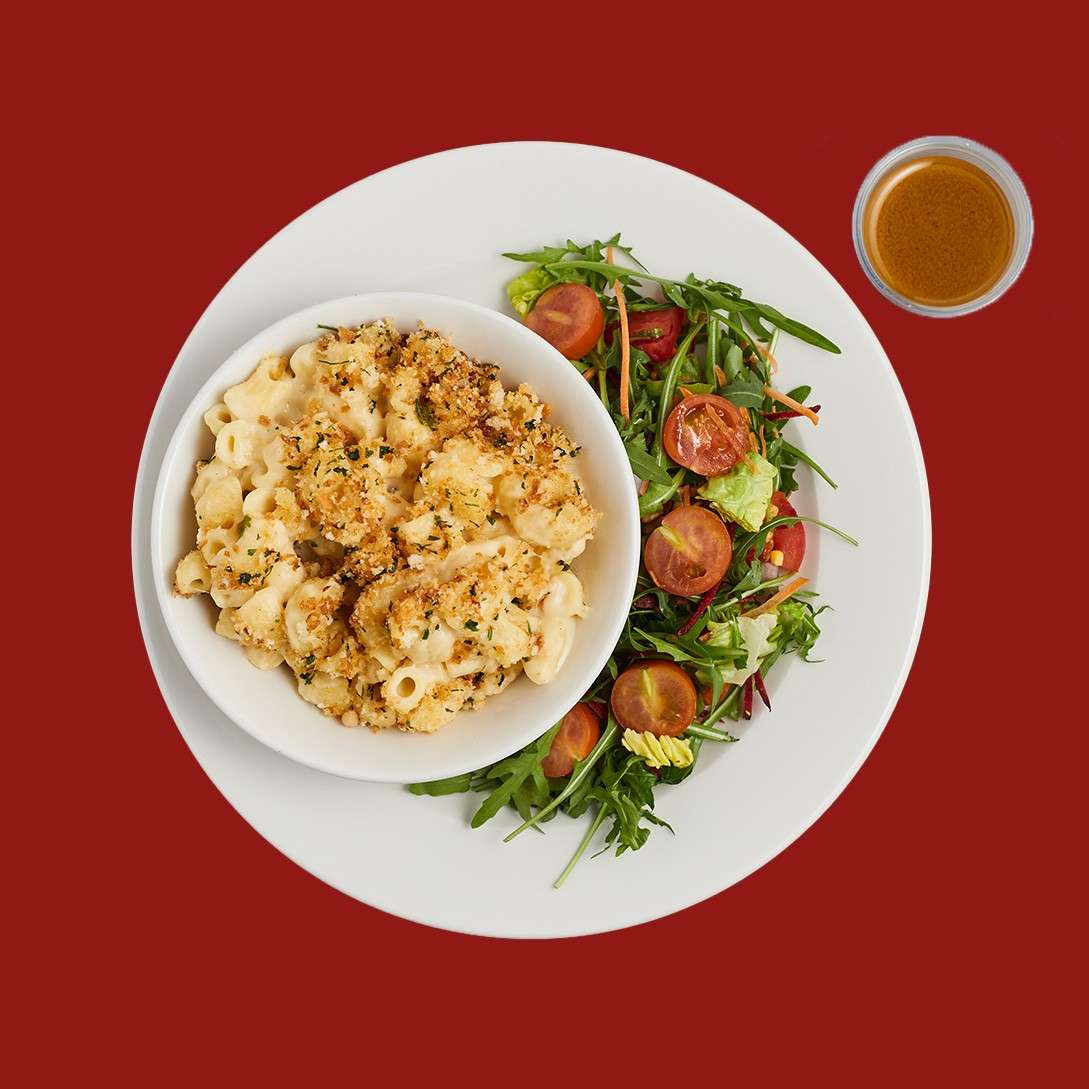 Macaroni and cheese in a bowl, topped with breadcrumbs; mixed green salad with cherry tomatoes alongside on a white plate; a small cup of dressing nearby on a red background.