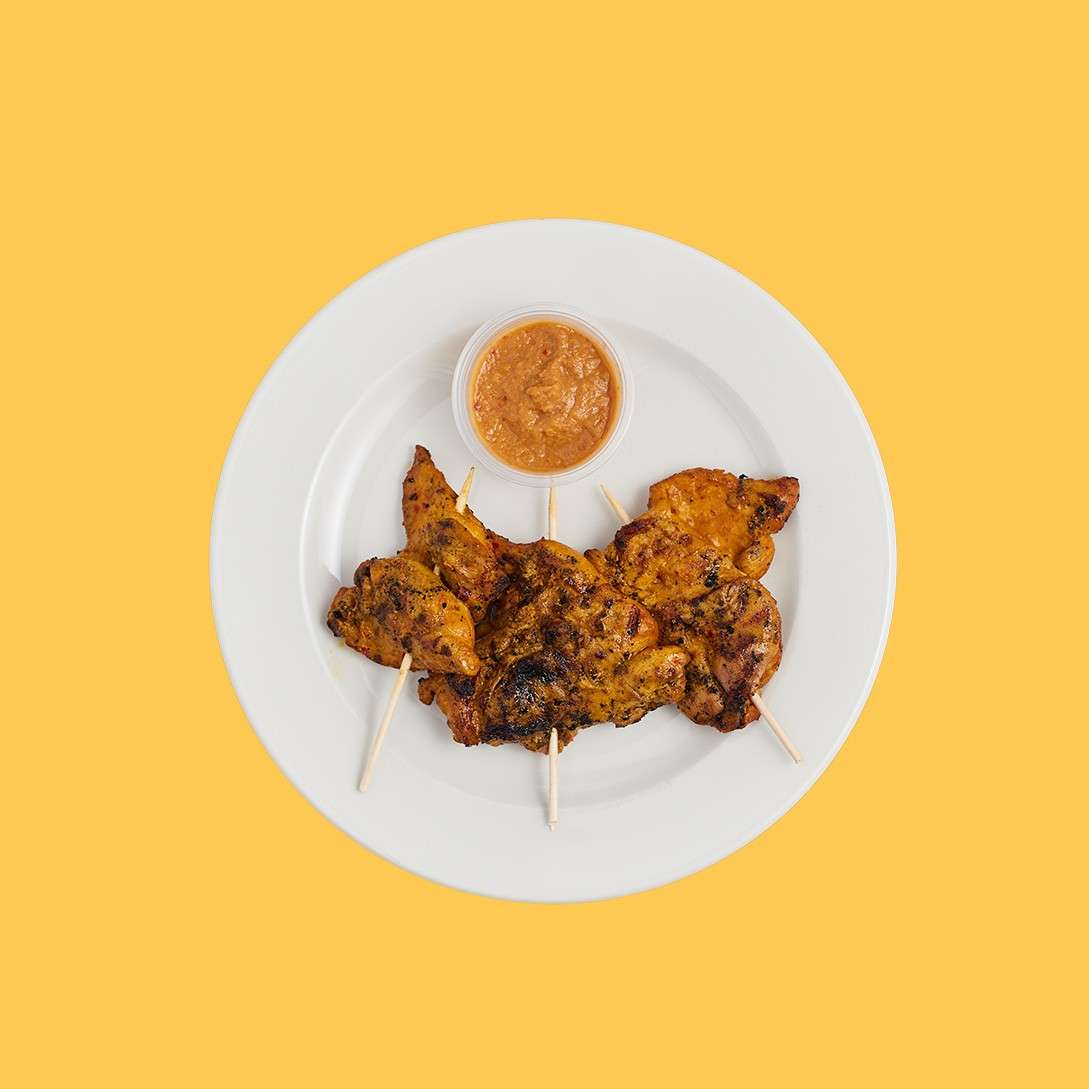 Grilled chicken skewers rest on a white plate, accompanied by a small cup of dipping sauce. Set against a yellow background, the skewers are charred and arranged with wooden sticks.