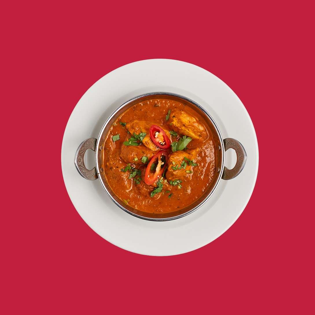 Bowl of chicken curry sits garnished with sliced red chili and cilantro, resting in a small metal handled dish on a white plate, centered against a solid red background.