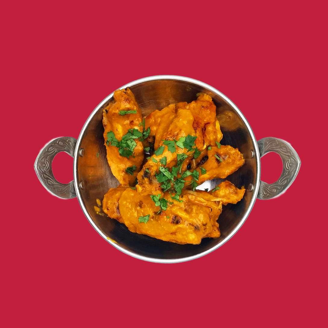 Sauced chicken pieces rest, garnished with chopped green herbs, in a round metal bowl with ornate handles, set against a solid red background.