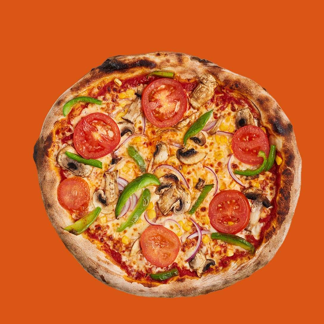 Pizza topped with tomato slices, green bell pepper, mushrooms, red onion, corn, and melted cheese sits centered, showing a charred crust, against a solid orange background.