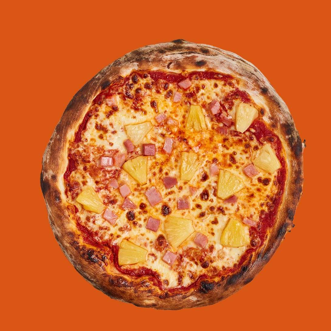 A round pizza topped with pineapple chunks and diced ham rests face-up, cheese melted over tomato sauce, charred crust visible, against a solid orange backdrop.