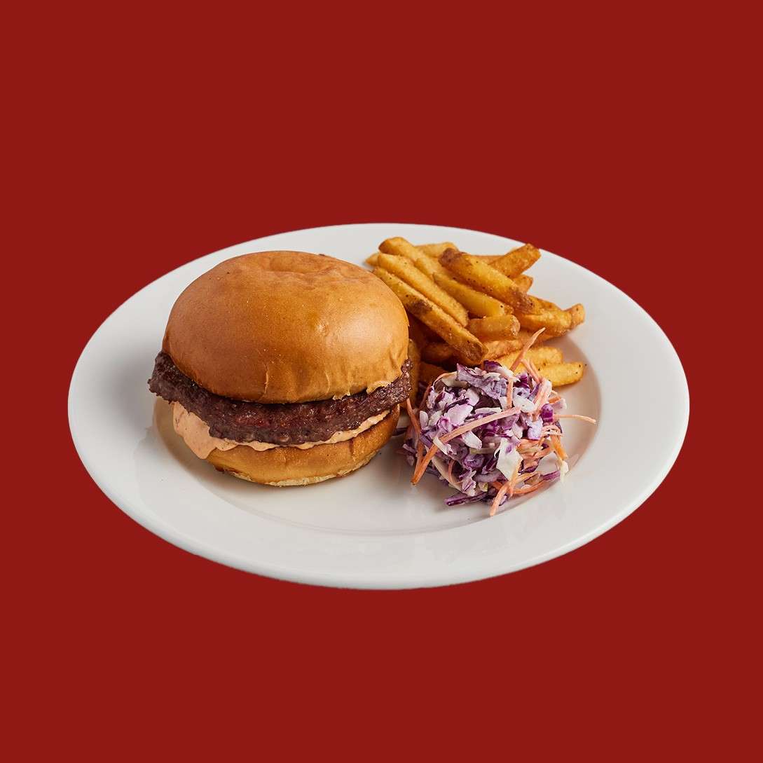 Burger rests on a white plate, accompanied by seasoned fries and purple cabbage slaw; context: isolated on a solid red background.