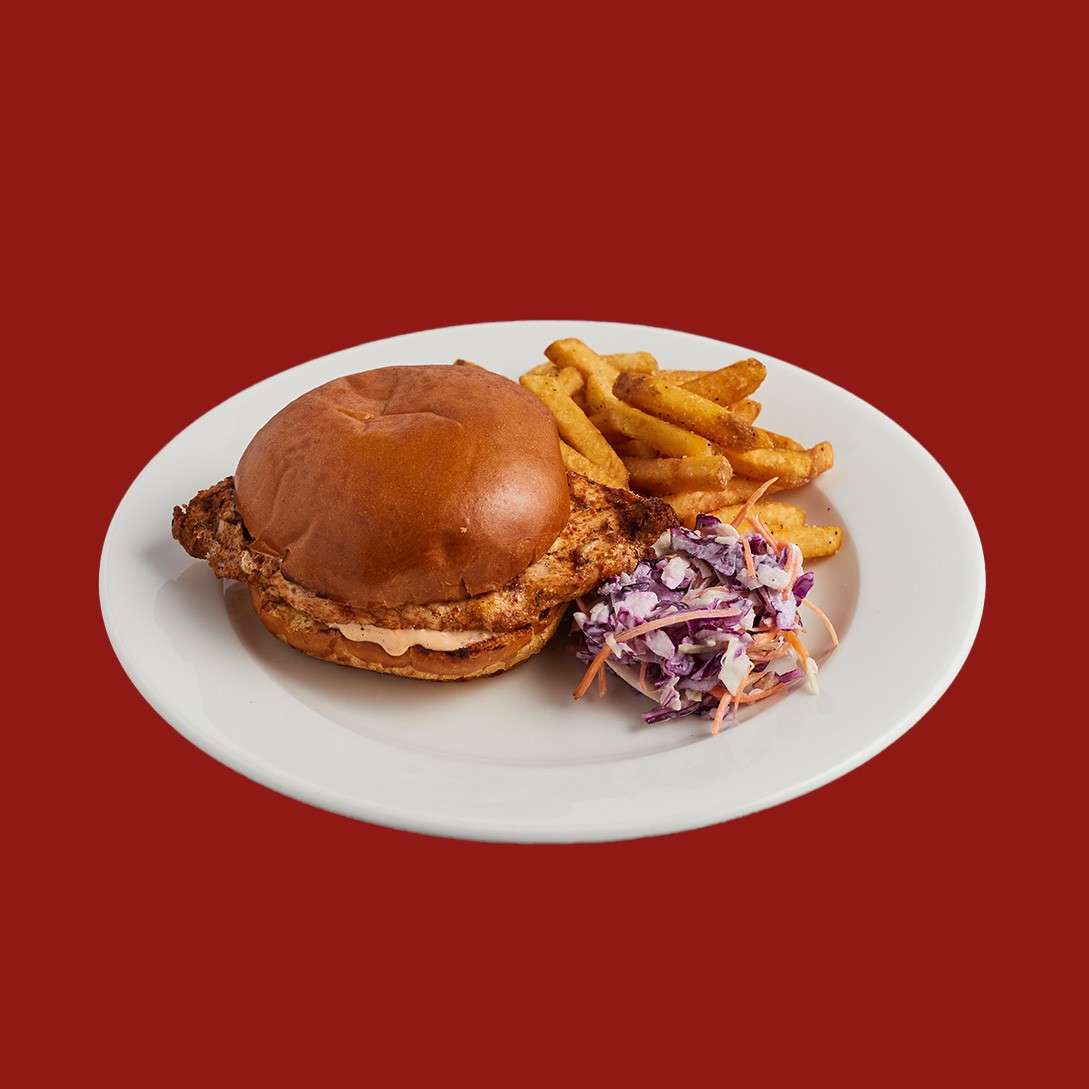 A grilled chicken sandwich rests on a white plate, paired with seasoned fries and purple coleslaw, set against a solid red background.