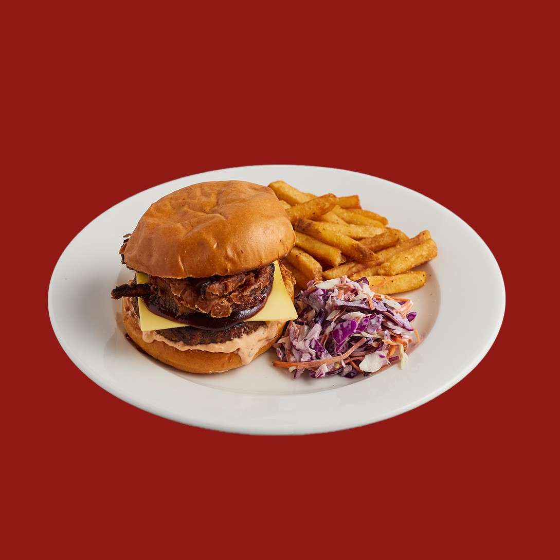 Cheeseburger with melting cheese and sauce rests on a white plate, accompanied by seasoned fries and purple-and-orange coleslaw, against a solid red background.