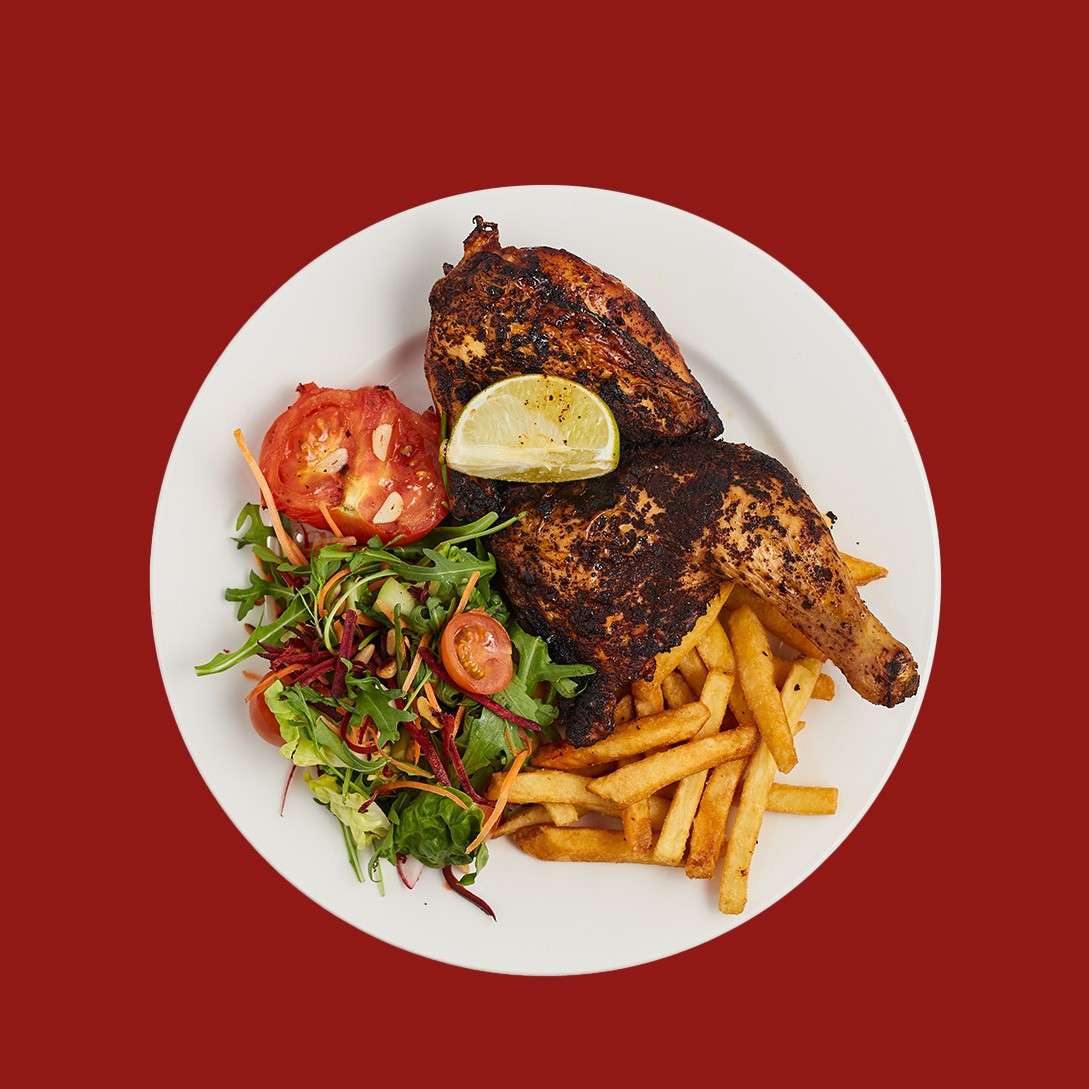 Grilled chicken quarter topped with a lime wedge rests on a white plate, served with golden fries and mixed salad, set against a solid dark red background.