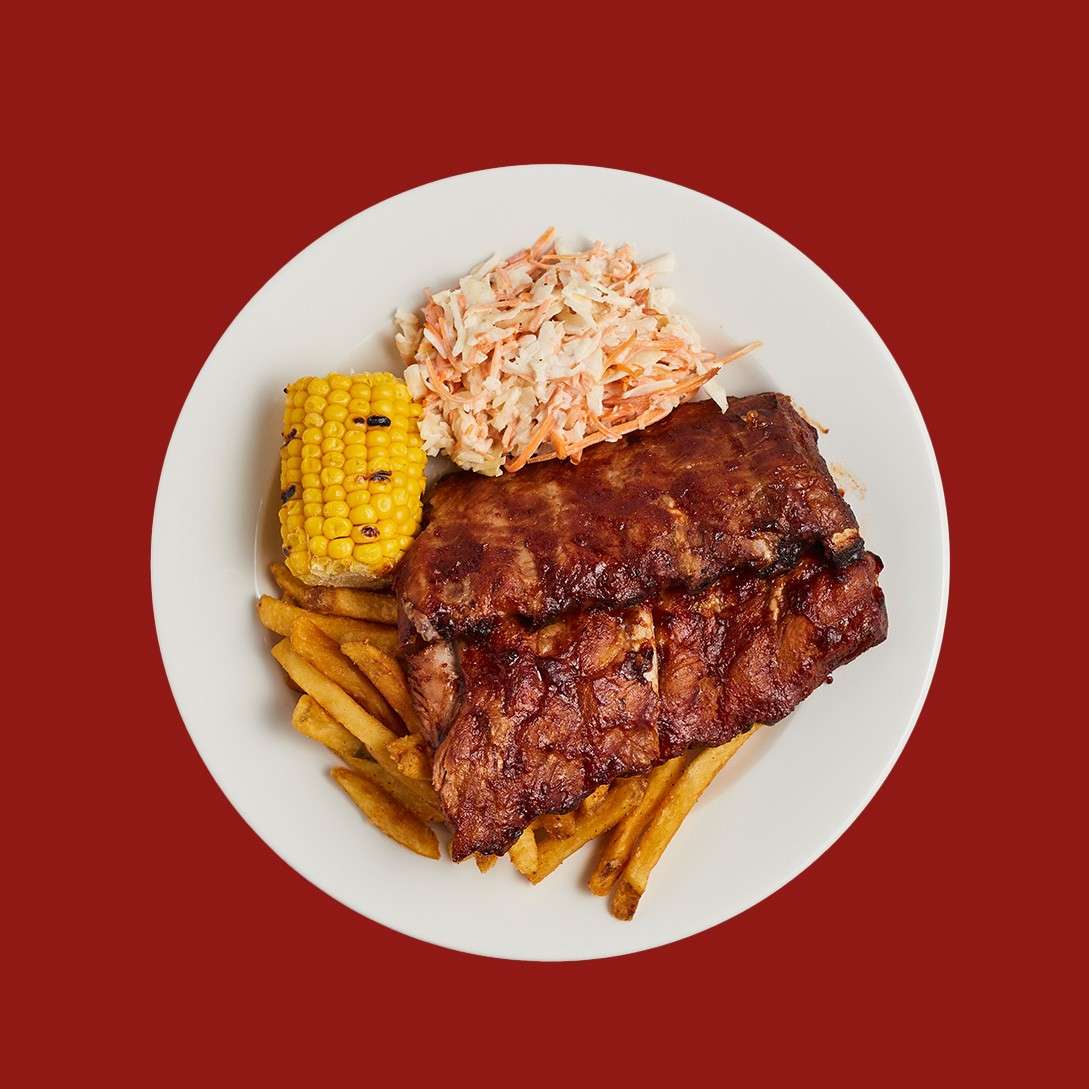 Barbecued ribs sit atop fries, accompanied by coleslaw and a grilled corn cob, on a white plate centered against a solid red background.