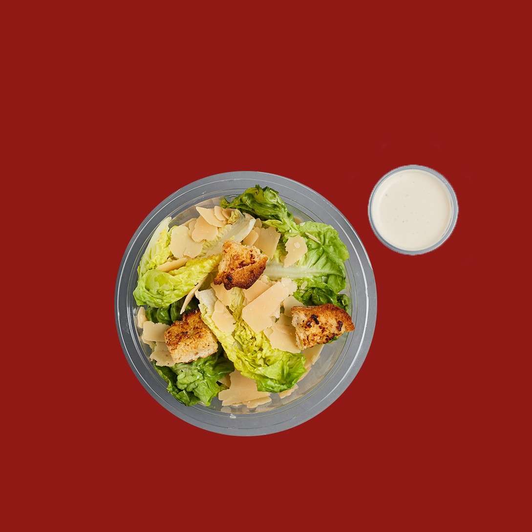 A bowl of Caesar salad rests, topped with lettuce, croutons, and shaved Parmesan; a small cup of creamy dressing sits beside it on a plain red background.