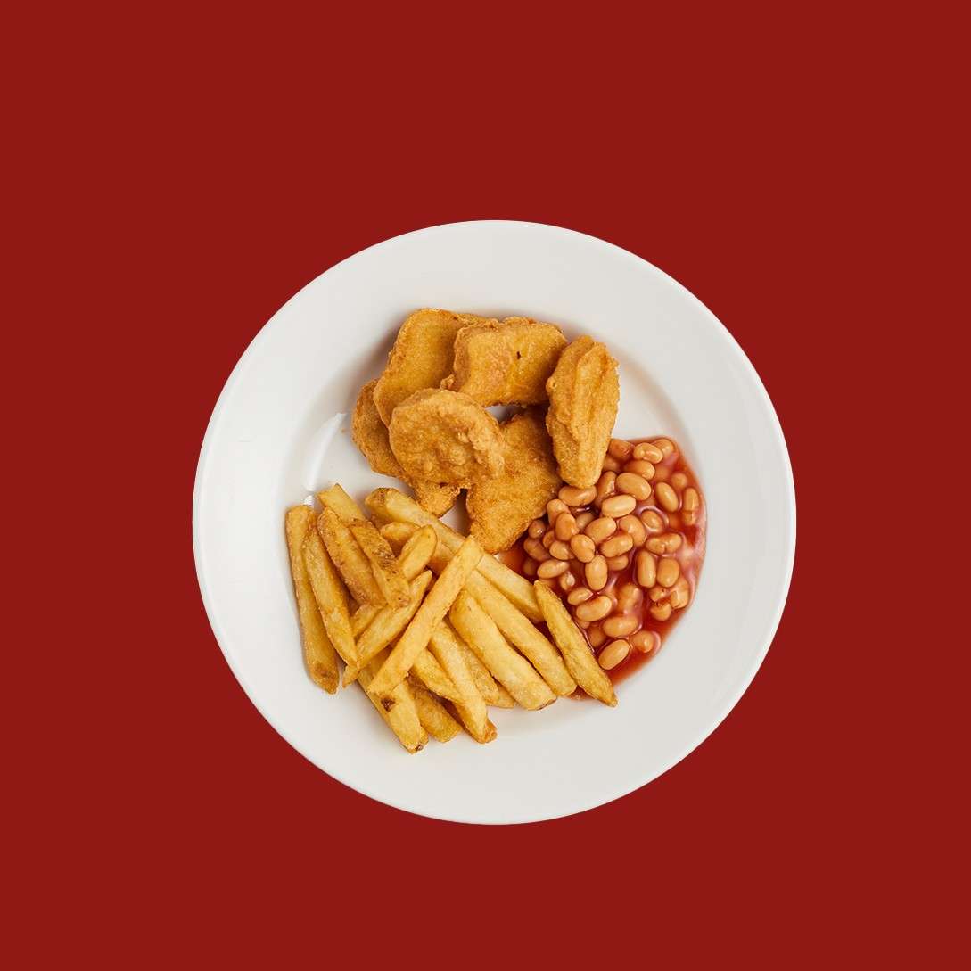 Chicken nuggets rest beside fries and baked beans on a white plate; viewed from above against a solid red background.