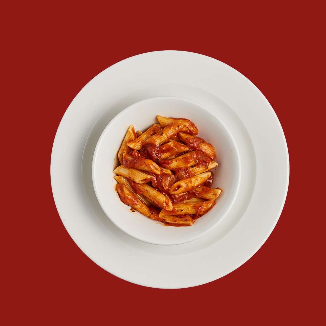 Penne pasta coated in tomato sauce sits in a small white bowl placed on a larger white plate against a solid dark red background.