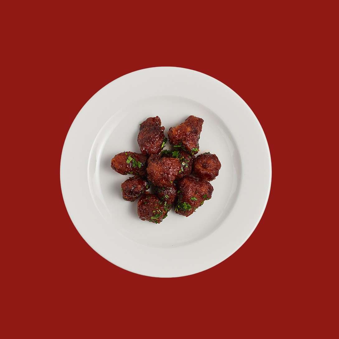 Sauced bite-sized fried pieces cluster together, glistening with dark red glaze and sprinkled with chopped herbs, resting on a white plate set against a solid red background.