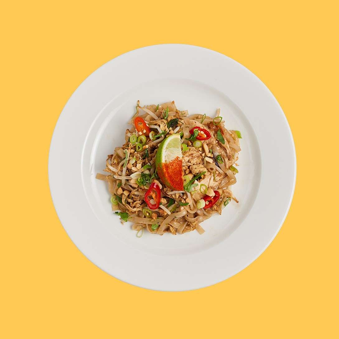 A white plate of Pad Thai sits centered, garnished with lime wedge, red chilies, scallions, peanuts, and bean sprouts, on a solid yellow background.