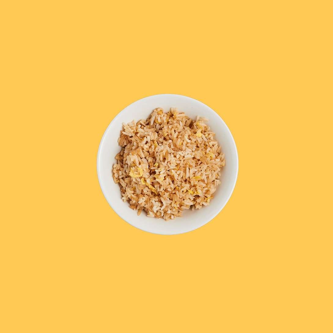 White bowl of fried rice rests centered, showing grains and egg pieces, on a plain mustard-yellow background.