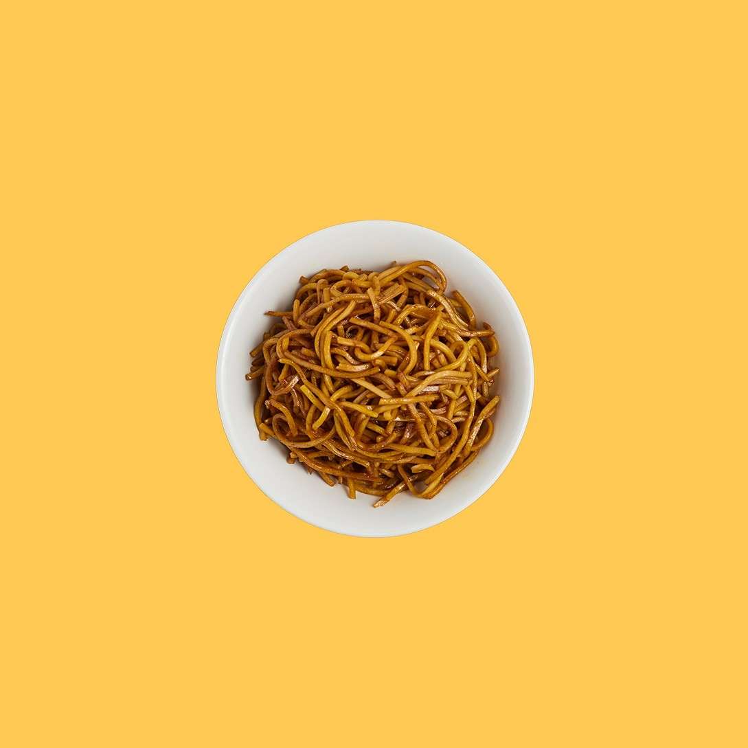 A white bowl of soy-sauce noodles sits centered, viewed from above, on a plain yellow background.