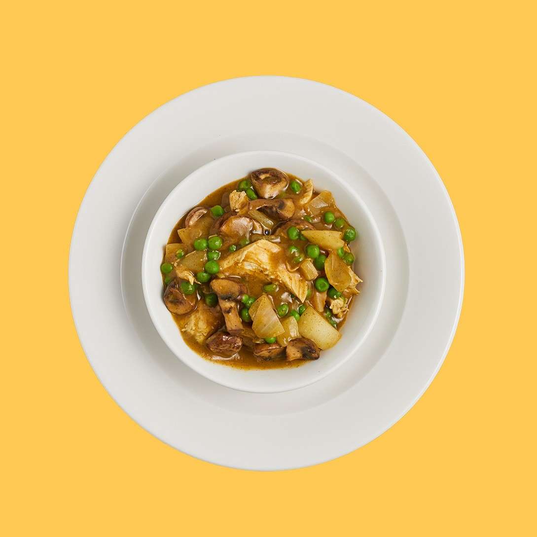 Bowl of chicken and vegetable stew sits in a white bowl, topped with peas, mushrooms, onions, and potatoes, resting on stacked white plates against a solid yellow background.