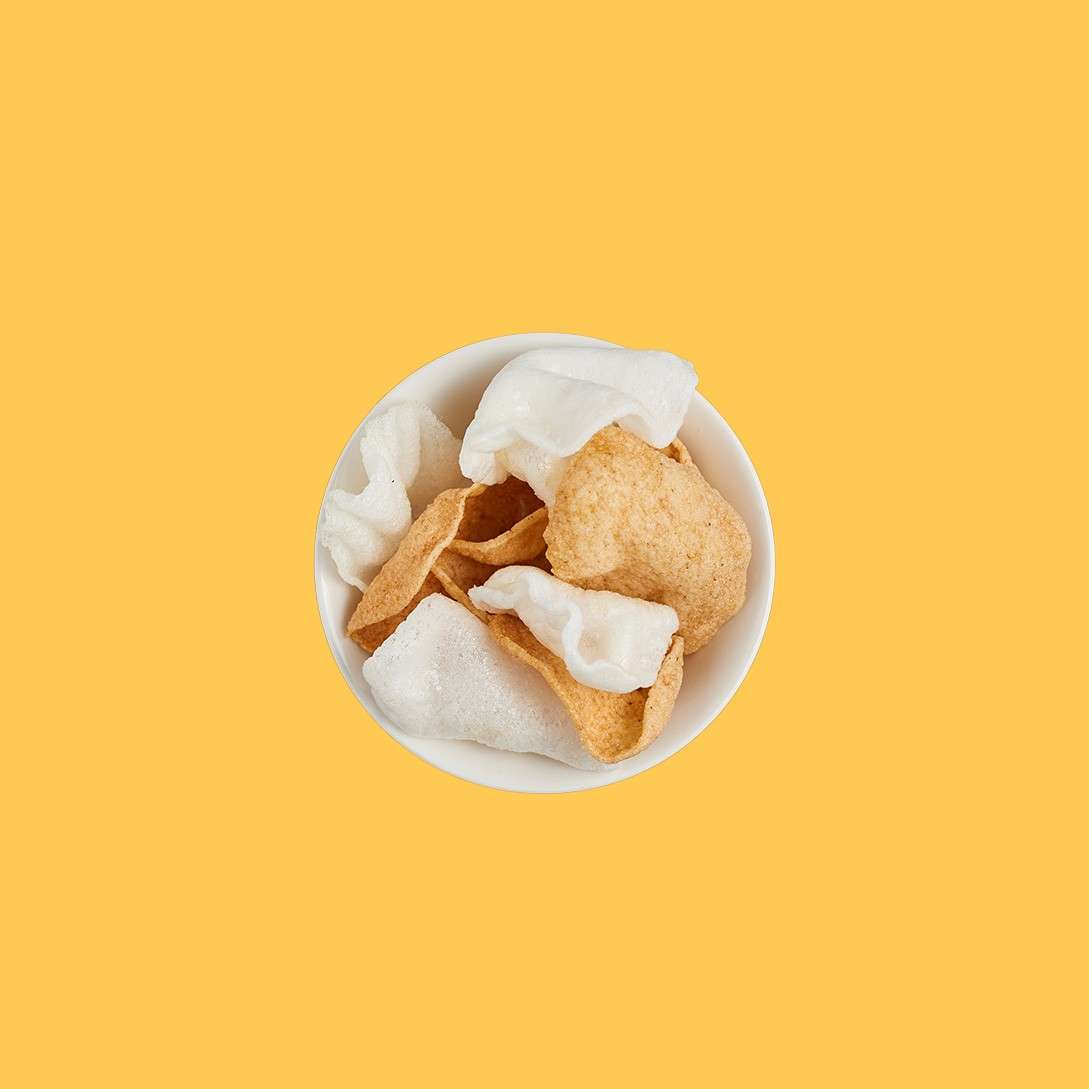Bowl of mixed crisps sits centered, showing white puffy crackers and tan chips, viewed from above. Set against a solid mustard-yellow background with ample empty space.