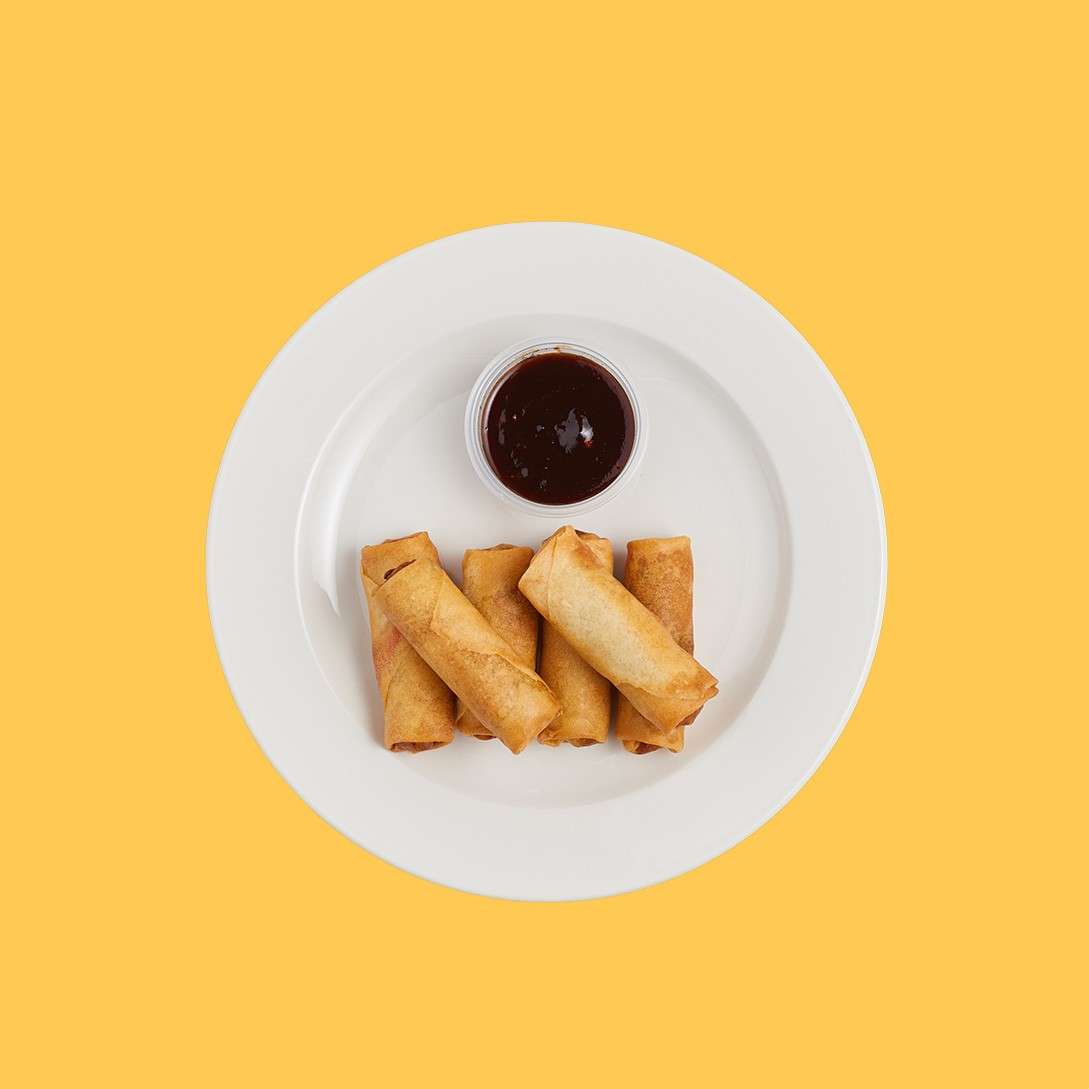 Four golden spring rolls rest beside a small cup of dark sauce on a white plate, viewed from above against a solid yellow background.