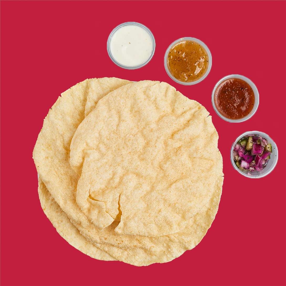 Crispy flatbread rounds stacked, accompanied by four small dipping cups: white sauce, amber chutney, red salsa, and chopped onion-cucumber mix, on a solid red background.