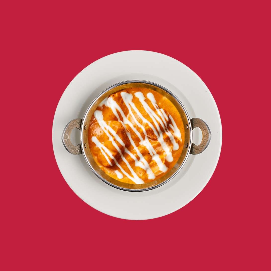 A small metal bowl of orange curry, drizzled with white sauce, rests on a white plate. The dish’s handles face outward, centered against a plain, solid red background.