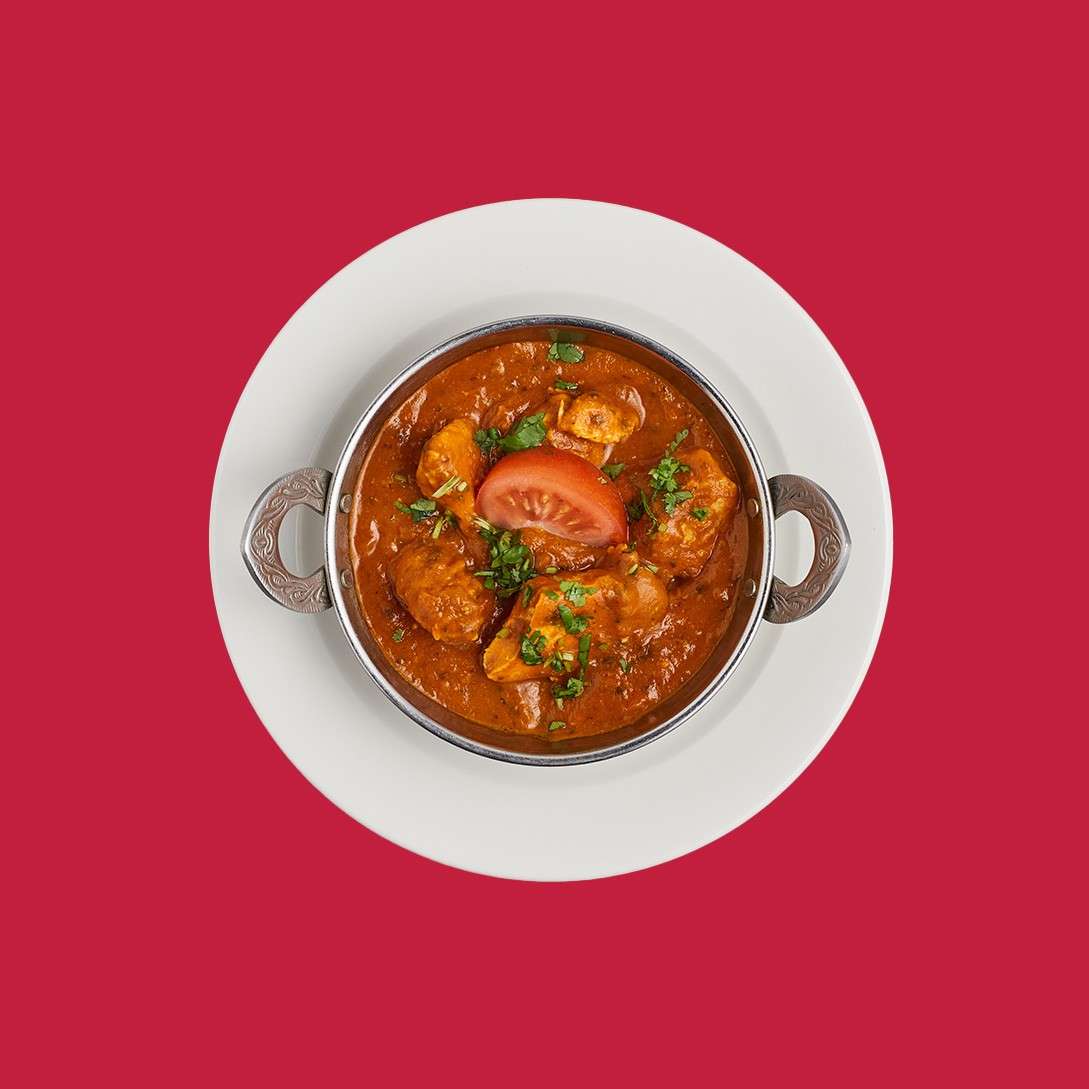 Chicken curry sits garnished with cilantro and a tomato wedge, in a handled metal bowl. Served on a white plate, overhead, against a solid red background.