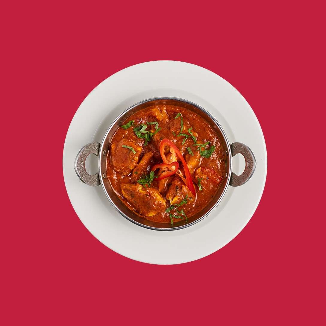 Metal handled bowl of red curry with chicken pieces, topped with sliced red chili and cilantro, sits on a white plate against a solid red background.