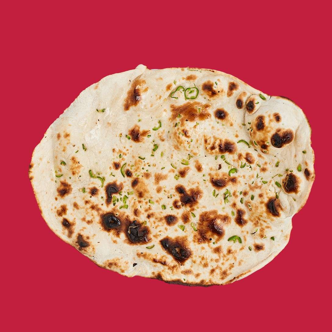 Flatbread rests, charred and blistered with scattered green herb flecks, showing uneven bubbles and browned spots. Context: isolated on a solid deep-red background.