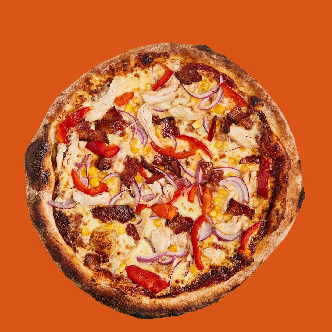Pizza topped with melted cheese, red onion, red pepper, corn, chicken pieces, and bacon sits centered, showing charred crust and scattered toppings, against a flat solid orange background.