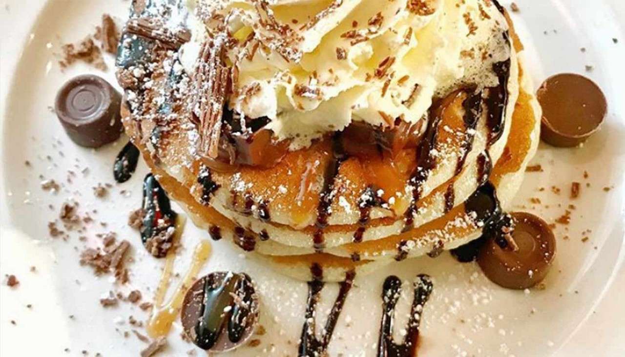 Stack of pancakes topped with whipped cream, chocolate shavings, and caramel, drizzled with chocolate sauce, sprinkled with powdered sugar; round chocolate candies surrounding, served on a white plate.