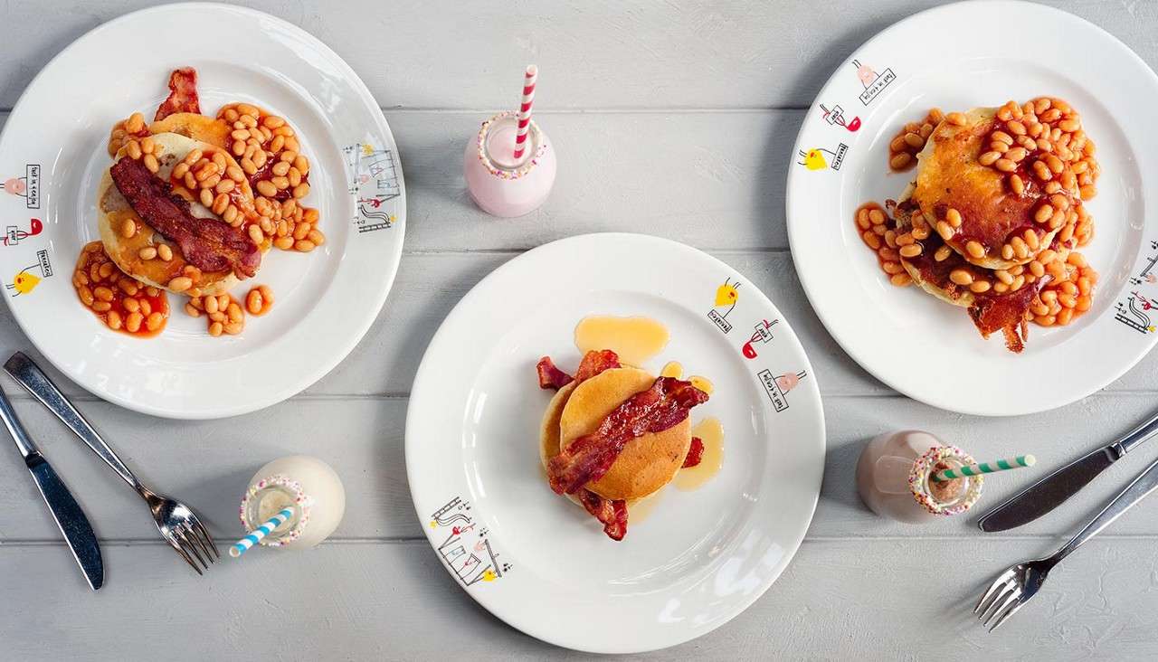 Pancakes topped with crispy bacon and baked beans sit on three white cartoon-decorated plates, accompanied by small milkshakes with striped straws and cutlery, on a light gray wooden table; plate text is illegible.