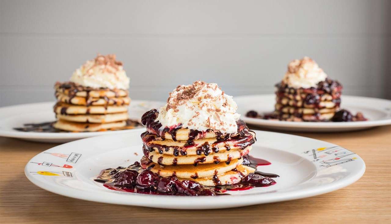 Stack of pancakes drips with berry compote and chocolate, topped with whipped cream and chocolate shavings; set on a decorated plate, with two similar pancake stacks blurred in the background.