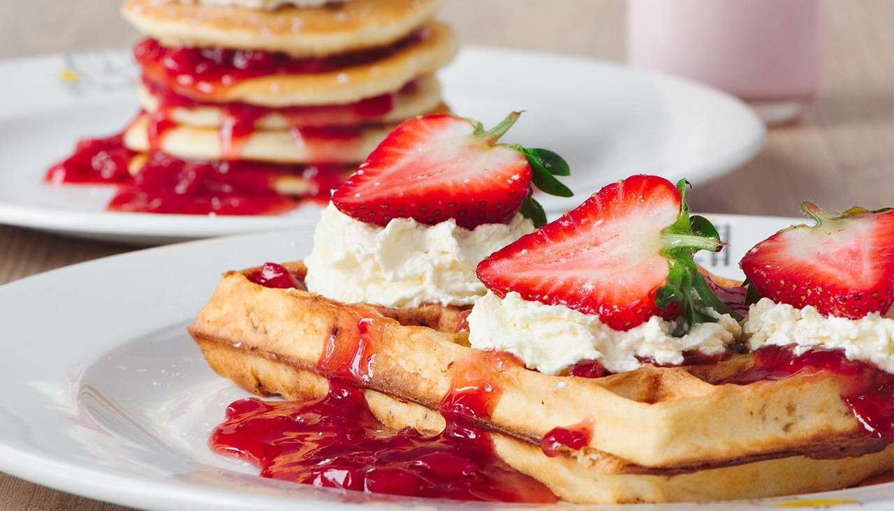 Waffles topped with whipped cream and halved strawberries, drizzled with red berry sauce, sit on a white plate; in background, a stack of pancakes with sauce and a pink drink.