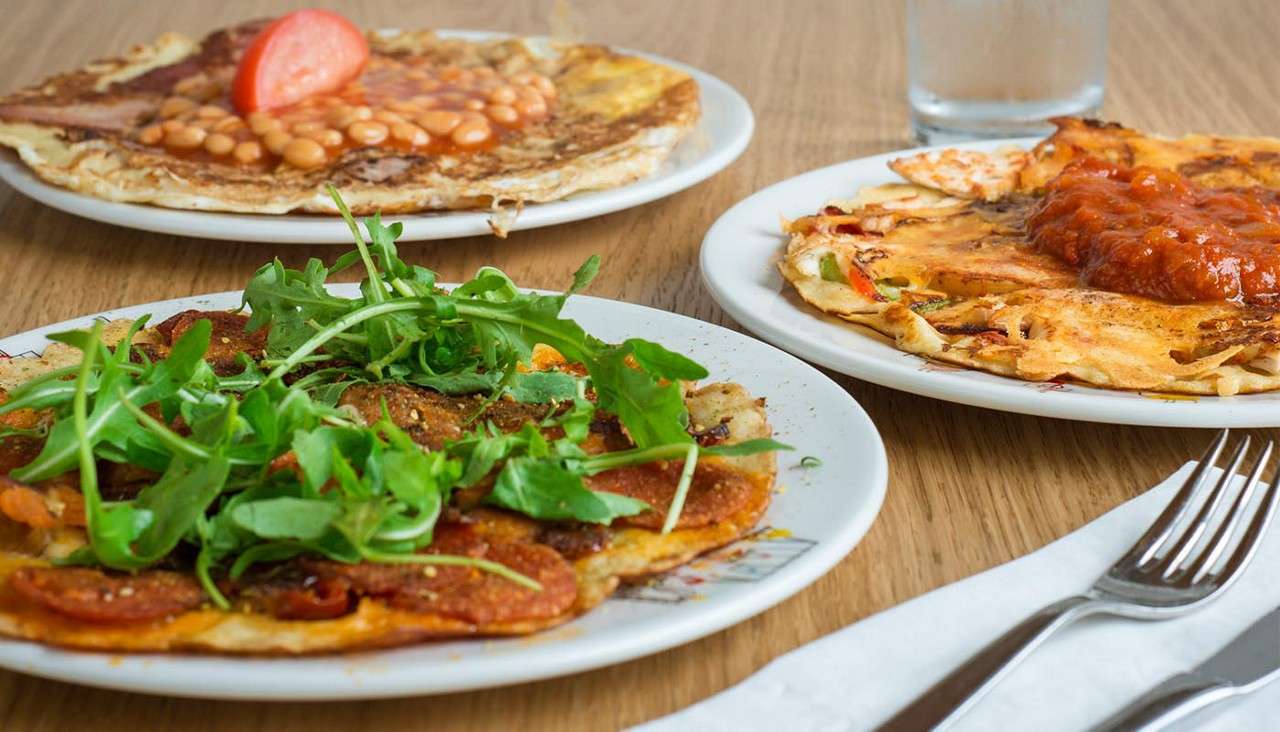 Three savory crepes sit on plates, topped with arugula and pepperoni, baked beans with tomato, and tomato sauce, resting on a wooden table beside cutlery, napkin, and a glass.