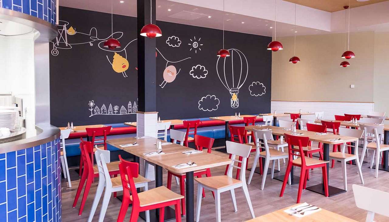 Colorful chairs and wooden tables sit arranged, awaiting diners; red pendant lamps hang above in a restaurant with chalkboard-style wall art: airplane, birds, hot-air balloon, clouds, and a small town.