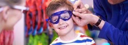 Boy wearing blue goggles—smiling as an adult adjusts the strap—in a brightly lit store or workshop, with colorful items blurred in the background.