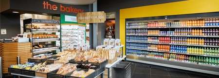 Pastries and loaves sit arranged on trays and shelves, while colorful bottled drinks line a refrigerated wall; context: a grocery store bakery section with a sign reading: the bakery.