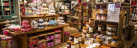Colorful soaps and bath products sit stacked and packaged on wooden tables and shelves, filling a cozy boutique store with assorted jars, candles, and gift boxes.