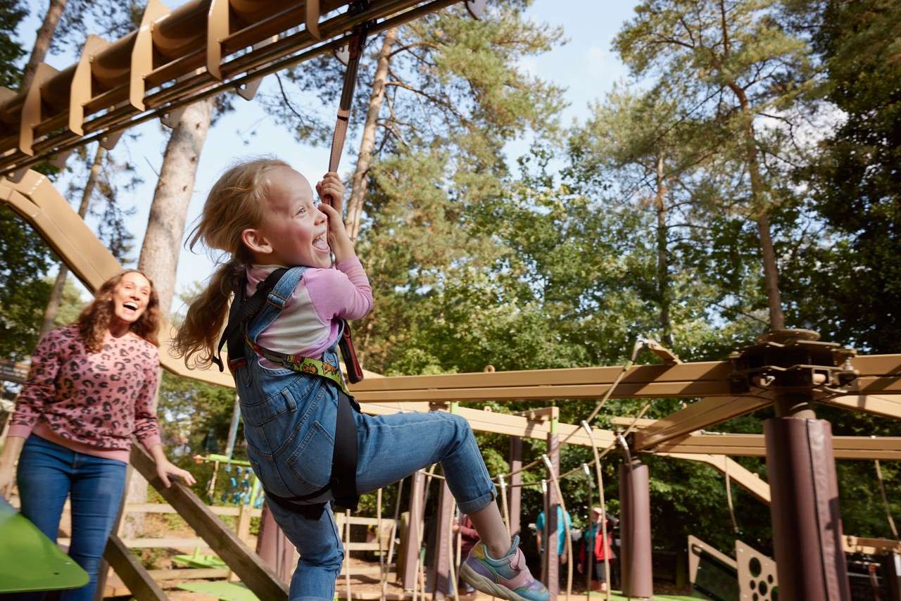 Child in denim overalls rides a suspended zip-line handle, smiling and gripping tightly; nearby adult watches excitedly. Wooden play structure and ropes sit among tall trees on a sunny day.