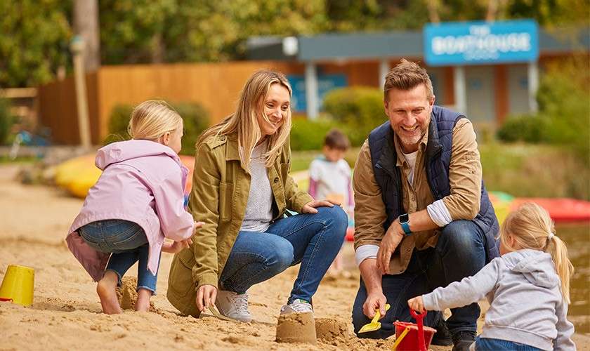 Parents and children build sandcastles with buckets and shovels, smiling together on a sandy lakeside beach; colorful kayaks and a building labeled “BOATHOUSE” appear in the background.