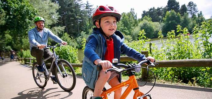 Child on an orange bicycle pedals ahead, wearing a red helmet; an adult follows on a bike. They ride along a paved path beside a wooden fence, trees, and water.