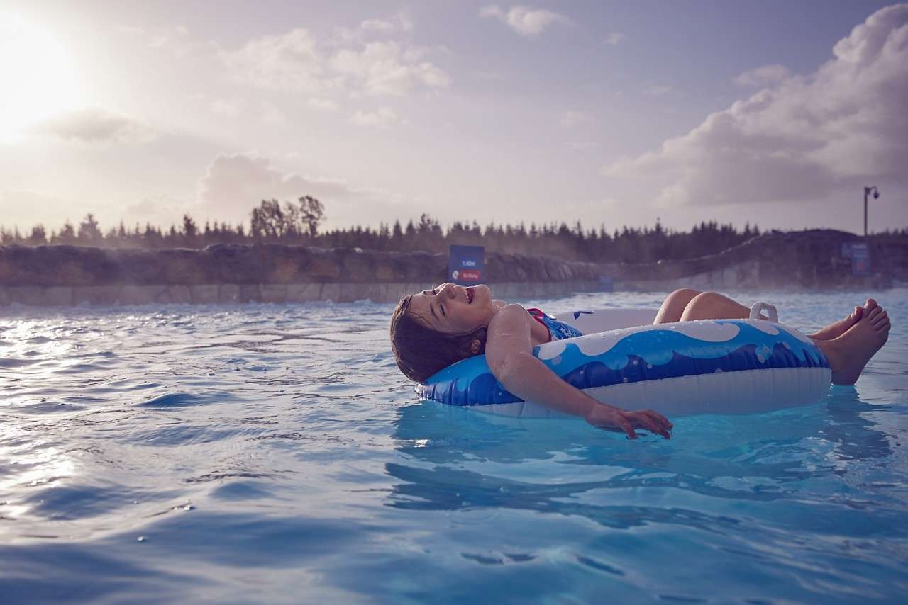 Child reclines on a blue inflatable tube, smiling and drifting. The tube floats in a calm outdoor pool, with misty water, rocky edging, distant trees, and a cloudy evening sky.