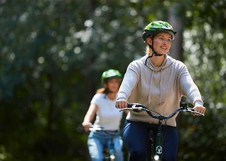 Two helmeted cyclists ride forward along a shaded forest path; the nearer rider in a beige hoodie leads, smiling and gripping handlebars, while another follows slightly out of focus among trees.