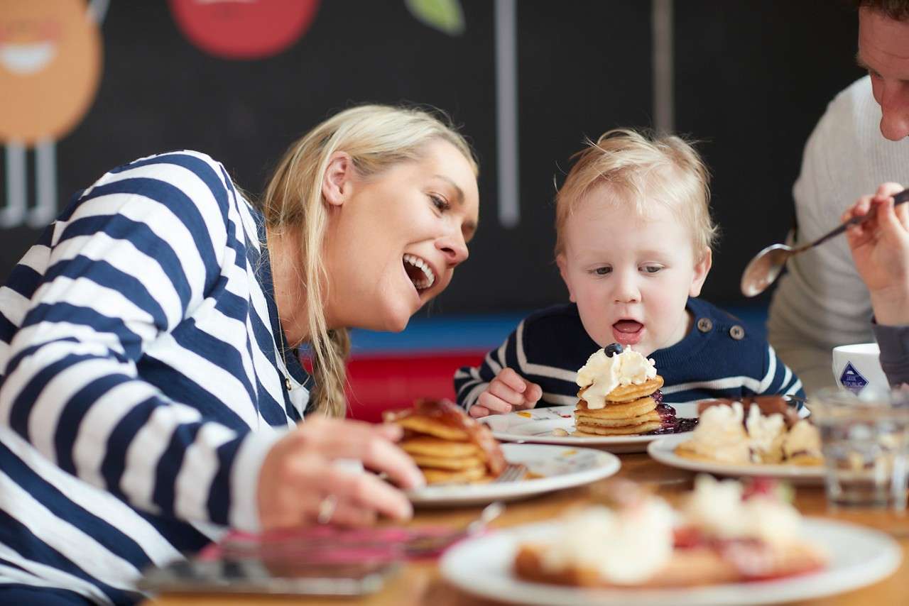 Two adults and two children laugh while eating at a round restaurant table; plates of salad, burgers, and drinks under warm lighting, with a toddler in a high chair.