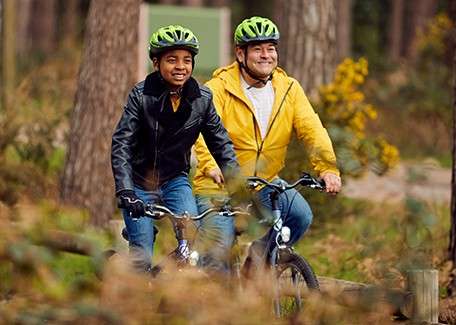 Two cyclists ride side by side, smiling, wearing green helmets. They pedal along a forest trail, surrounded by tall trees and shrubs; one wears yellow, the other black.