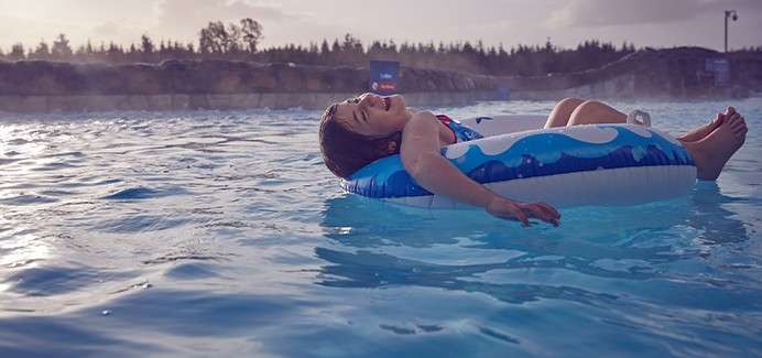 Child floats reclining on a blue inflatable tube, smiling upward; in an outdoor pool with gentle ripples, a rocky edge, distant trees, and hazy evening light.