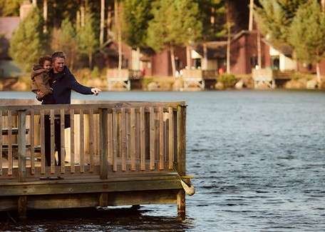 Adult holding a child points across the water, standing on a wooden dock; calm lake surrounds them, with pine trees and rustic cabins lining the opposite shore in soft light.