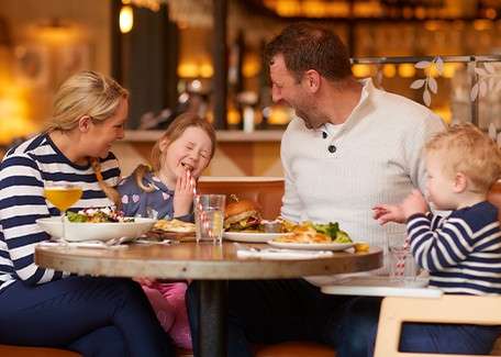 Family of four shares a meal, laughing together; older child giggles, hand over mouth, toddler in a high chair. Plates of salad, burger, and fries sit on a restaurant table.