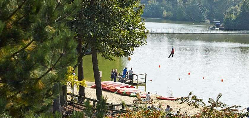 Person zip-lines over a calm lake, gliding toward shore; on the sandy beach below, kayaks lie beside a small dock while people watch and trees frame the recreational park setting.