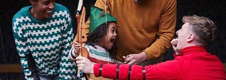 Child with green hat holds a wooden bow and laughs, as two adults guide and another cheers, during a friendly archery lesson in a dim, sheltered outdoor area.